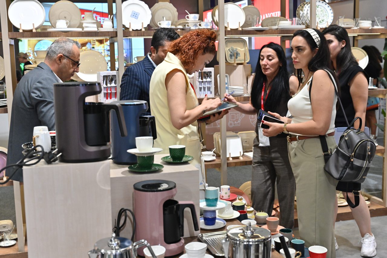 Bir grup ziyaretçi, sergi standında oturmuş, birinin konuşmasını dikkatle dinliyor.